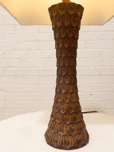 Load image into Gallery viewer, A vintage-style table lamp with a sculpted, flared base featuring a repeating honeycomb or textured geometric pattern in dark brown. The lamp is lit, casting a warm glow from a cream-colored shade, set against a white painted brick wall.