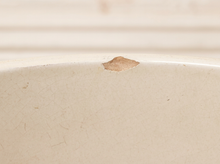 Load image into Gallery viewer, Close-up of a cream-colored ceramic surface with a small chip in the glaze near the rim.