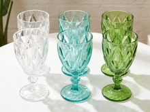 Load image into Gallery viewer, Six cut-glass goblets in white, aqua, and green, arranged on a white table against a light brick wall background.