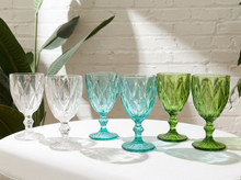 Load image into Gallery viewer, Six cut-glass goblets in white, clear, turquoise, and green shades, arranged on a white table against a white brick wall with leafy plants in the background.