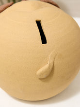 Load image into Gallery viewer, A close-up of a round, matte beige ceramic piggy bank with a vertical slot on top and a small protruding tail detail, shown on a white surface.