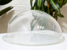 Load image into Gallery viewer, A clear, dome-shaped plastic food cover or domed lid with a flat base, shown on a white surface with a blurred background of white brick wall and green plants.