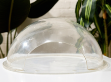 Load image into Gallery viewer, A clear acrylic dome cover with a semi-circular arch design, shown resting on a white surface with blurred green plants in the background.