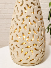 Load image into Gallery viewer, A teardrop-shaped ceramic lamp with an organic cutout pattern, glowing with warm light from within, set against a white brick wall background.