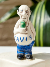 Load image into Gallery viewer, A ceramic figurine of a bald, wrinkled man holding a green mug, with the name 'GAVIN' written in blue on his belly. The figurine is standing on a white surface with a blurred background of green leaves.