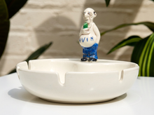 Load image into Gallery viewer, A white ceramic ashtray with a sculpted figure of a bald man wearing blue pants and holding a green object, with the name 'EVAN' written on his belly, displayed on a white surface with a blurred background.