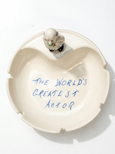 Load image into Gallery viewer, An off-white ceramic ashtray with a small figurine of an actor at the rim, featuring the handwritten blue text 'THE WORLD'S GREATEST ACTOR' in the center, set against a plain white background.