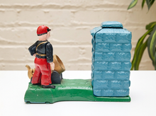 Load image into Gallery viewer, A vintage cast iron decorative block featuring a firefighter standing next to a blue brick chimney on a green base, set against a white brick wall background.