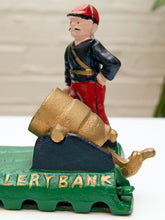 Load image into Gallery viewer, A cast iron novelty bank featuring a soldier figurine on a green base with gold lettering that reads 'CURRENCY BANK'. The soldier is wearing a red hat, black jacket, and red trousers, with a gold barrel on a dark blue platform.