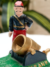 Load image into Gallery viewer, A small decorative figurine of a mustached man in a red hat and dark blue outfit with gold trim, standing behind a gold cannon mounted on a green base. The background is softly blurred with green leaves and a neutral surface.