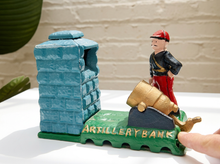 Load image into Gallery viewer, A small decorative cast-iron artillery bank featuring a blue brick tower, a green base with gold lettering, a gold cannon, and a soldier figurine, shown on a white surface with a finger pointing at it.