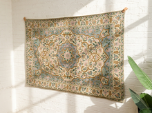 Load image into Gallery viewer, A large vintage-style area rug with an intricate floral and medallion pattern in muted blue, beige, pink, and green tones, hanging on a white brick wall with sunlight casting shadows.
