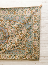 Load image into Gallery viewer, A vintage-style oriental rug with a floral medallion pattern in muted shades of beige, dusty blue, pink, and olive green, hanging against a white painted brick wall.