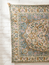 Load image into Gallery viewer, An ornate vintage-style area rug with a floral medallion pattern, hanging against a white painted brick wall.