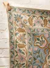 Load image into Gallery viewer, A close-up of a woven textile or rug hanging from a wooden peg against a white painted brick wall. The pattern features cream, dusty blue, pink, and olive green floral and vine motifs.