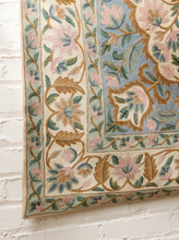 Load image into Gallery viewer, A close-up of an ornate area rug with a floral pattern, featuring a light blue field, pink and green floral motifs, and a beige border, shown hanging against a white brick wall.