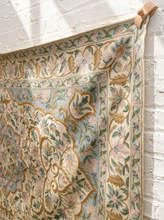 Load image into Gallery viewer, An ornate vintage-style tapestry with a floral and scroll pattern in muted pink, sage green, and gold on a cream background, hanging from a wooden dowel against a white brick wall.