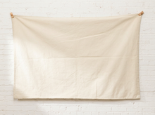 Load image into Gallery viewer, A large cream-colored linen fabric panel or tablecloth hanging flat against a white painted brick wall, secured at the corners with wooden pegs.