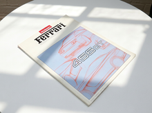Load image into Gallery viewer, A printed Ferrari magazine or booklet lies open on a white round table, showing a light blue interior spread with red line drawings of a car and the large number 456 GT. The visible brand is Ferrari.