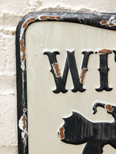 Load image into Gallery viewer, A close-up of a distressed vintage-style metal sign with a cream background, black lettering, and a black border showing peeling white paint and exposed brown rust. The sign is mounted against a white painted brick wall.