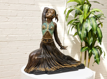 Load image into Gallery viewer, An ornate bronze-finished sculpture of a dancer in a flowing dress with turquoise and gold patina details, displayed on a white pedestal against a white brick wall next to a green plant.