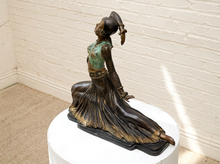 Load image into Gallery viewer, A bronze-finished sculpture of a kneeling dancer with a flowing skirt, displayed on a white pedestal against a white brick wall.