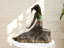 Load image into Gallery viewer, A bronze-finished sculpture of a dancer in a flowing dress, displayed on a white pedestal against a white brick wall with soft leaf shadows.