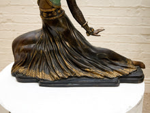 Load image into Gallery viewer, A bronze-finished sculpture of a seated female figure with flowing draped fabric and gold decorative accents, displayed on a white pedestal against a light brick wall.