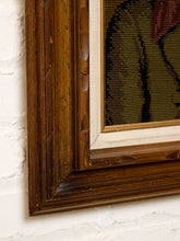 Load image into Gallery viewer, Close-up of a framed artwork showing a dark brown carved wooden frame with a cream mat and a needlepoint canvas depicting a figure in olive green, black, and burgundy.