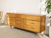 Load image into Gallery viewer, Mid-century modern wooden dresser with six drawers, tapered legs, and brass handles, positioned in a room with a white brick wall and a potted plant.