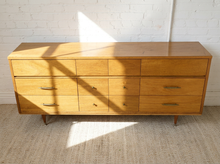 Load image into Gallery viewer, A mid-century modern wooden dresser with six drawers, brass handles, and tapered legs, positioned against a white brick wall on a textured beige rug with dramatic sunlight casting shadows.