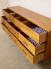 Load image into Gallery viewer, A mid-century modern wooden storage cabinet with multiple drawers and open cubbies, shown with several drawers pulled open to reveal patterned fabric lining. The cabinet is positioned on a textured beige carpet against a white brick wall.
