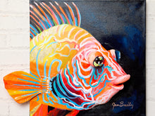 Load image into Gallery viewer, A colorful painted fish artwork on a square canvas, mounted against a white painted brick wall. The fish illustration features bright yellow, orange, pink, red, and blue tones on a dark navy background, with the artist signature 'Jean Bradley' visible in the lower right.