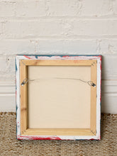 Load image into Gallery viewer, The back view of a square framed canvas print, showing a light wood frame with a distressed, paint-splattered border in white, red, and blue, mounted on a beige woven rug against a white brick wall.
