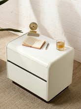 Load image into Gallery viewer, A modern cream-colored two-drawer nightstand with a glossy finish, styled with an open book, a small clock, a pen, and a glass of whiskey on top, set against a white brick wall and beige rug.