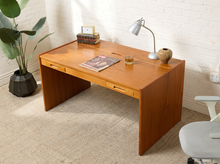Load image into Gallery viewer, Image 3 of 22: A mid-century modern wooden writing desk with a warm honey finish, styled in a room with a white brick wall, beige carpet, and a potted plant.