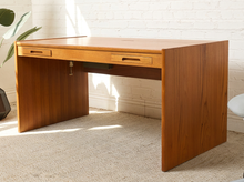 Load image into Gallery viewer, Image 5 of 22: A mid-century modern wooden writing desk with two drawers, shown in a room setting with a white brick wall and beige carpet.