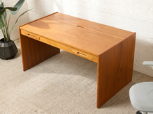 Load image into Gallery viewer, Image 6 of 22: A mid-century modern wooden desk with a warm honey finish, featuring two drawers on the front and a cable grommet cutout on the top surface, set in a room with a white brick wall and beige carpet.