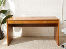 Load image into Gallery viewer, Image 4 of 22: A mid-century modern wooden writing desk with two drawers, positioned against a white brick wall on a textured beige rug, with a potted plant on the left.