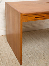 Load image into Gallery viewer, Image 9 of 22: A close-up of a mid-century modern wooden desk with a warm honey finish, featuring a single drawer with a recessed pull handle, set on a textured beige rug against a white brick wall.