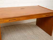 Load image into Gallery viewer, Image 10 of 22: A mid-century modern wooden desk with a warm honey finish, featuring a pull-out drawer on the left, a pull-out drawer on the right, and a drop-down side panel. The desk is set on a textured beige rug against a white brick wall.