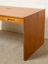 Load image into Gallery viewer, Image 12 of 22: A mid-century modern wooden writing desk with a warm honey-toned finish, featuring a single drawer with a pull handle, set against a white brick wall and textured beige carpet.
