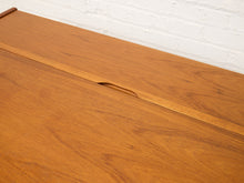 Load image into Gallery viewer, Image 14 of 22: Close-up of a mid-century modern wooden table with a warm honey-brown finish, featuring a recessed handle detail and a white painted brick wall in the background.