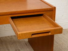 Load image into Gallery viewer, Image 18 of 22: Close-up of a mid-century modern wooden desk with a warm honey-toned finish, showing an open drawer with a recessed pull handle, set against a white brick wall and textured rug.