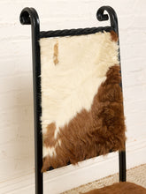 Load image into Gallery viewer, Image 7 of 15: A black metal chair with a cowhide upholstered backrest, featuring a natural brown and cream patchwork pattern, set against a white brick wall.