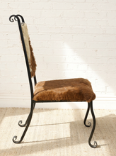 Load image into Gallery viewer, Image 5 of 15: A rustic side chair with a black wrought-iron scroll frame and a brown and beige animal-hide seat and backrest, set against a white brick wall on a light woven rug.