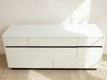 Load image into Gallery viewer, Image 5 of 18: A modern white wooden dresser with six drawers and minimalist black horizontal handles, positioned against a white brick wall on a light beige rug.