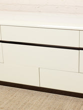 Load image into Gallery viewer, Image 7 of 18: Close-up of a cream-colored dresser with a glossy finish, featuring a dark wood accent strip running horizontally between drawers, set against a white brick wall and textured beige carpet.