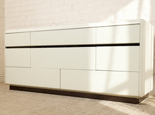 Load image into Gallery viewer, Image 11 of 18: A modern six-drawer dresser in glossy off-white with a dark wood base, positioned against a white brick wall with soft window light casting shadows.