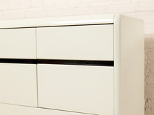 Load image into Gallery viewer, Image 12 of 18: Close-up of a cream-colored wooden dresser with a black horizontal handle, set against a white brick wall background.